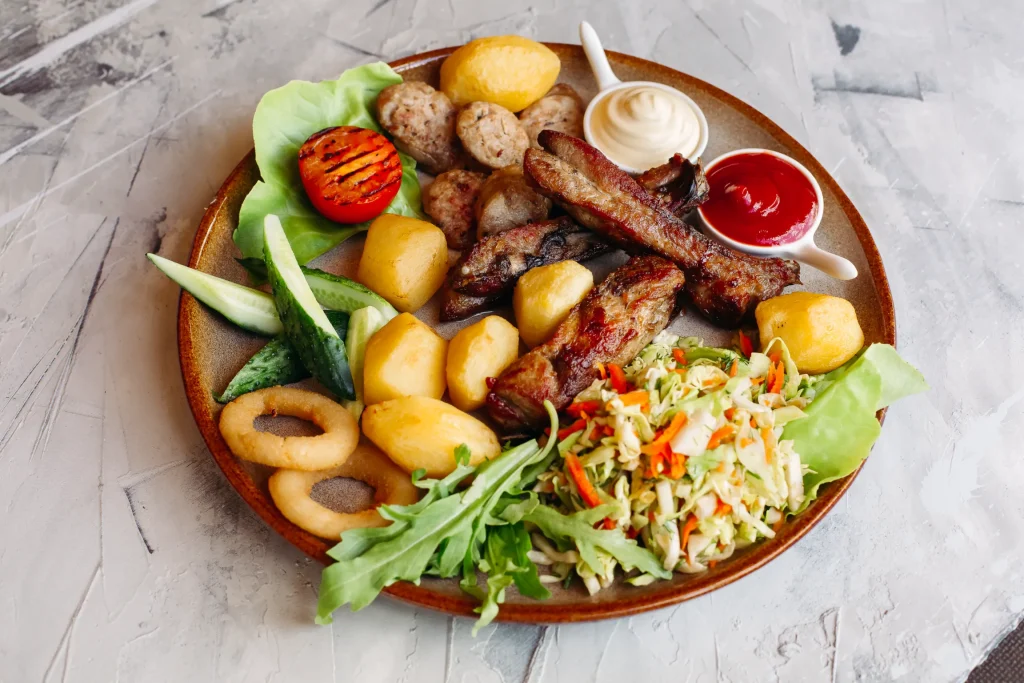 Here is a suggested alt text description for the current image: "Top-down view of a hearty meal on a wooden platter featuring grilled ribs, roasted potatoes, coleslaw, fresh salad greens with cucumber and a grilled tomato half, onion rings, and two small dipping bowls with red and white sauces, set against a textured grey background.