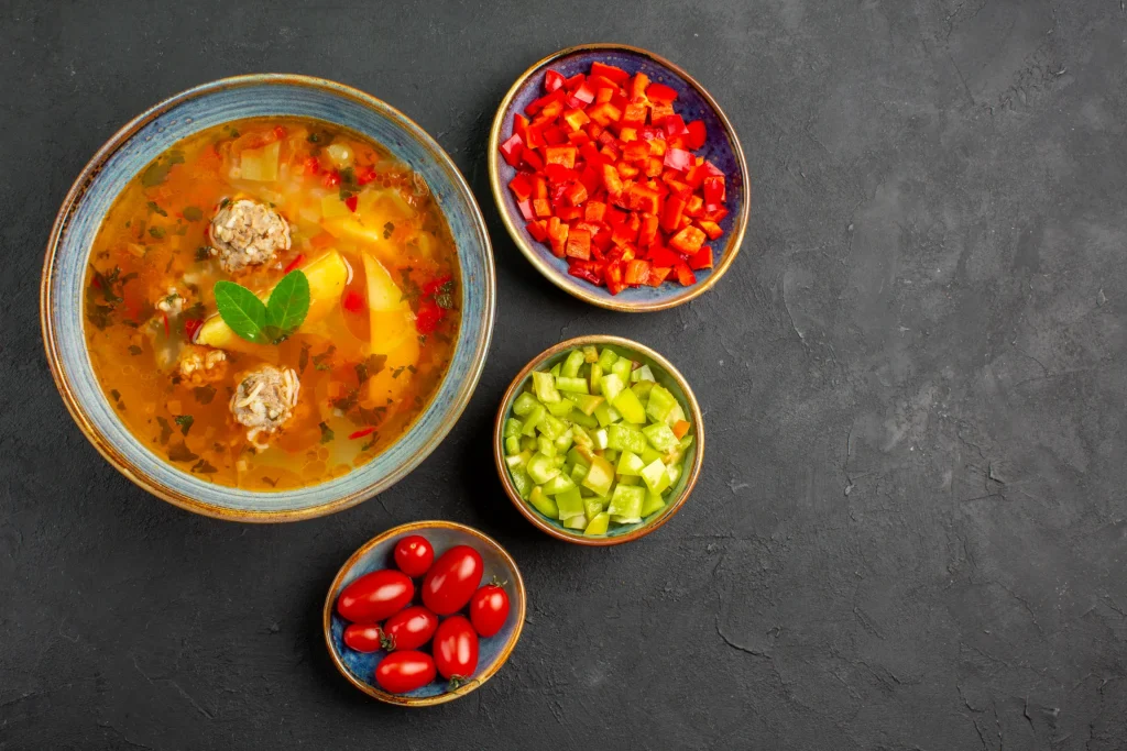 Top-down view of a bowl of hearty meat and vegetable soup garnished with fresh herbs, accompanied by three small bowls containing diced red peppers, diced green peppers, and cherry tomatoes, all arranged on a dark, textured background.
